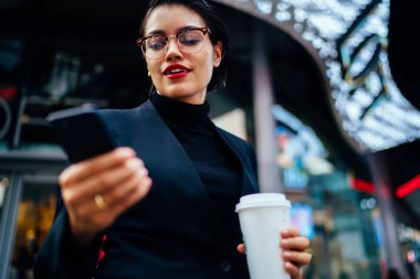 Gülümseyen iş kadını zarif giyim kahve tutan smartphone cep telefonu ile e-posta kutusunu kontrol içinde açık havada, başarılı kadın girişimci için online bankacılık stree üzerinde gezinme sırasında uygulama kullanarak gitmek