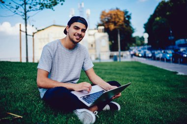 Dışarıda çimenlerin üzerinde oturan ve netbook üzerinde araştırma online yapma dinlenme neşeli hipster adam portresi