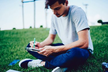 Genç öğrenci yeşil çimenlerin üzerine oturmuş defter denetim listesi yapma konsantre