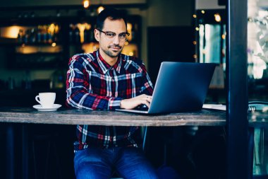 Optik Gözlük kameraya uzaktan modern dijital dizüstü bilgisayarda çalışırken arıyorum genç sakallı işadamı portresi. Yakışıklı erkek kafede oturan netbook klavye metin yazarak