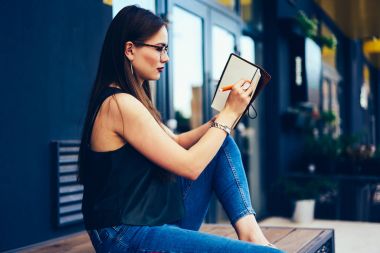 Düşünceli çekici kadın blogger şık Eyewear Notepad için kişisel web sitesi yayında yeni yaratıcı fikirler aşağı yazma. Dalgın hippi kız dışarıda otururken bilgi kaydetme