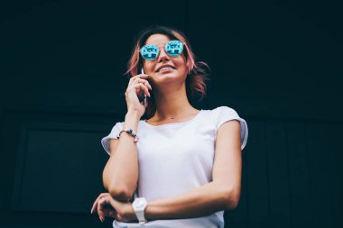 Şık mavi güneş gözlüklü genç başarılı Tasarımcısı görünümünün altındaki sahte alanı için smartphone aygıt ayakta promosyon siyah arka plan üzerine konuşurken kısa mesaj ile beyaz t-shirt giymiş