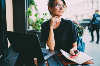 Genç çekici Grafik Tasarımcısı'nda uzağa bakarak ve çalışmalarında uygulamak için yaratıcı fikirler düşünmeye gözlükler. Şirin kız öğrencinin Not Defteri'nde Sokağı'nda otururken denetim listesi yapma