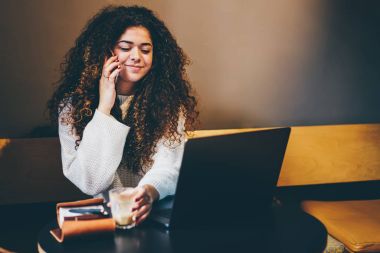 Arkadaş grubu telefonu mobil konuşmayı kıvırcık saçlı genç olumlu grafik tasarımcı. Mutlu sevimli kadın kişi Smartphone'da kafede dizüstü bilgisayarda uzaktan çalışma sırasında söz