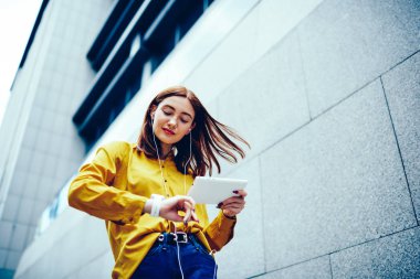 Kulaklık üzerinden favori şarkı listesi tadını çıkarırken şık kol saati zamanında kontrol rüzgarlı saçlı hoş yaratıcı kadın blogger. Sevimli genç kadın yaz yürüyüş sırasında modern aygıtla yeniden
