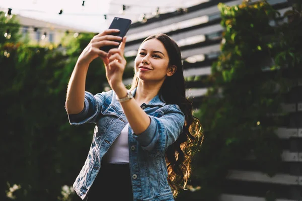 ブルネットの髪をフロント カメラ目線と現代のスマート フォンのデバイスの Selfies を作ると魅力的な女性 スタイリッシュなデニム ジャケット電話屋外で立っての撮影で笑顔の若い女性 — ストック写真