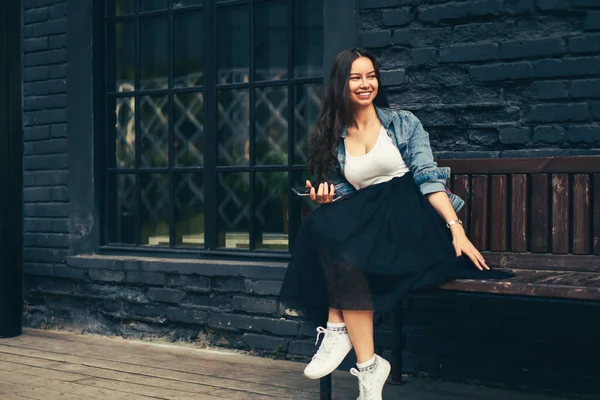 Beautiful cheerful young woman enjoying recreation time on wooden bench outdoors in urban setting.Positive female looking away while sitting near black house.Promotional background for advertising