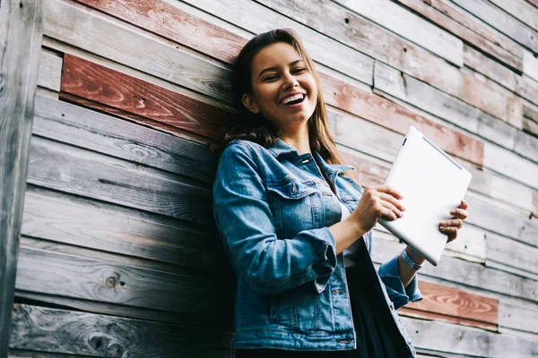 Half length portrait of positive young woman with modern tablet in hands smiling at camera.Funny hipster girl dressed in denim jacket holding touch pad while standing on promotional background