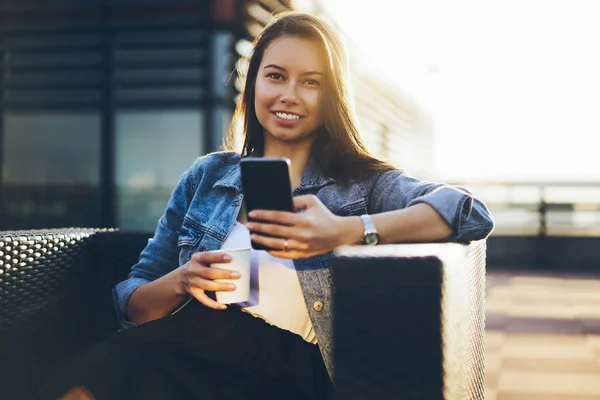 陽気な内気な少女は現代のスマート フォン デバイス上で友人に Sms メッセージを送信するデニム ジャケットに身を包んだ 肯定的なブロガーの屋外カフェに座って美味しいコーヒーを楽しむ携帯電話でオンライン チャット — ストック写真