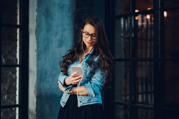 Charming stylish hipster girl in trendy eyeglasses listening favourite music in earphones connected to smartphone.Gorgeous female searching new songs on internet website for playlist on cellular