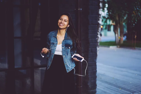 カメラの笑顔しながら都会の通りに散歩中に現代の電話デバイスは 音楽を聴くに接続されているデジタル イヤホンで陽気な若い女性の肖像画の長さ半分 — ストック写真