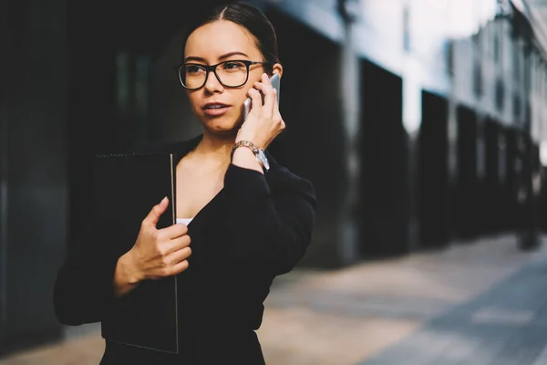 Confident young woman lawyer in optical eyeglasses talking with business partners via modern smartphone while standing outdoors and holding folder in hands.Copy space area for financial information