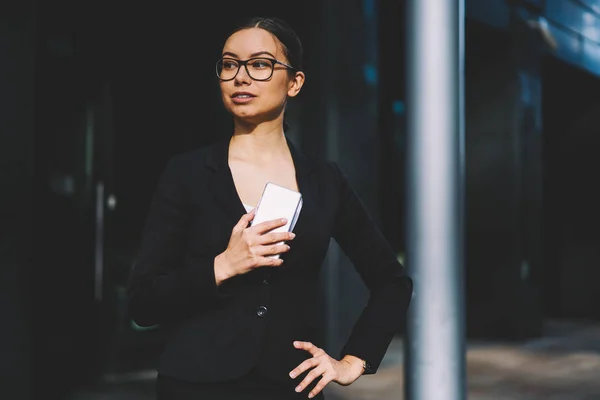 资深女性经济学家身着时尚的黑色西装和光学眼镜手持现代数字手机在手和看着 而站在办公大楼的宣传背景 — 图库照片