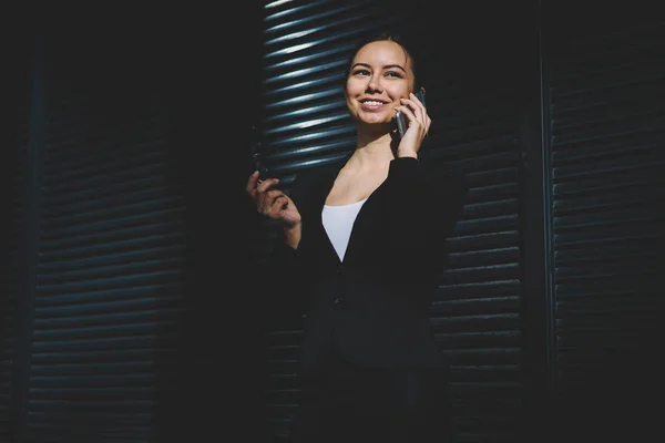 Prosperous female financial director dressed in elegant black suit holding eyeglasses in hand and talking with colleague on mobile phone standing near copy space area for your business information