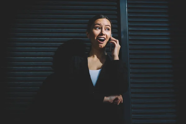 Shocked young woman executive manager dressed in black elegant apparel surprised with information during phone conversation standing in break near publicity area for your business advertising
