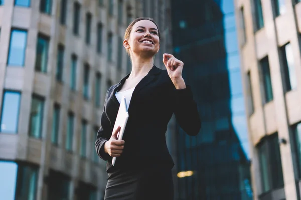 Happy businesswoman dressed in formal wear smiling and strolling outdoors in urban setting.Cheerful successful proud ceo holding digital tablet in hands standing on office promotional background