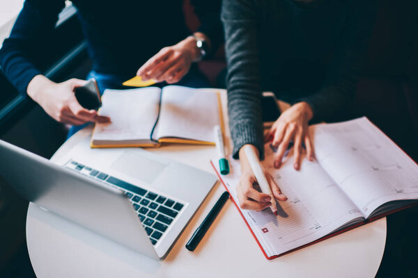 Cropped image of woman's hands recording information to notepad using marker while sitting at table indoors.Female creating working business plan while male colleague checking information in textbook