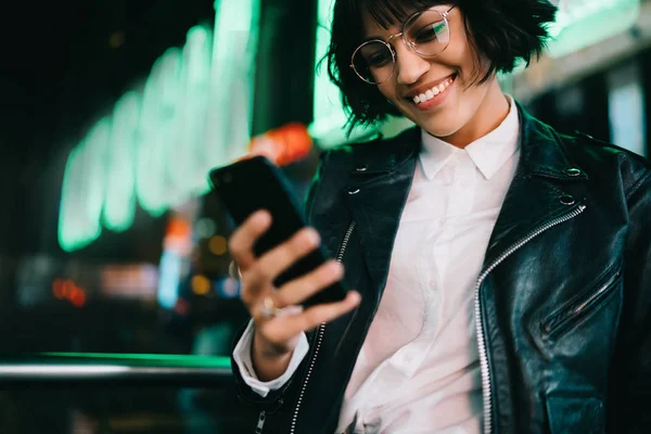Smiling hipster girl happy about getting message from friend checking mail on phone standing on urban background.