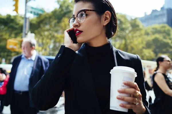 Confident businesswoman with red lips talking on phone walking on crowded city street, serious female entrepreneur in elegant formal wear having mobile conversation holding mock-up coffee cup