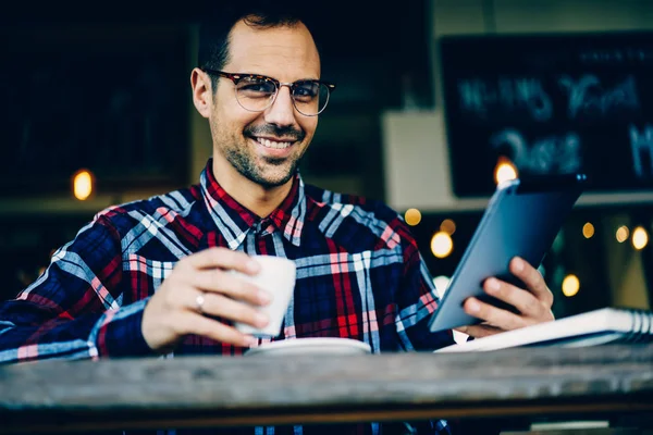 Cropped image of handsome bearded young man looking at camera ad smiling while holding cup of tasty coffee in hand and updating application on digital tablet via free 4G high speed internet connection