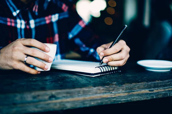 Cropped image of male's hands holding pen and writing down text information in notebook sitting indoors in cafe interior.Wooden table with cup of tasty coffee and notepad for noting some records