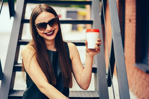 Half length portrait of cheerful brunette woman in stylish sunglasses having fun during strolling at street with coffee cup.Happy cute hipster girl looking at camera while sitting at urban setting