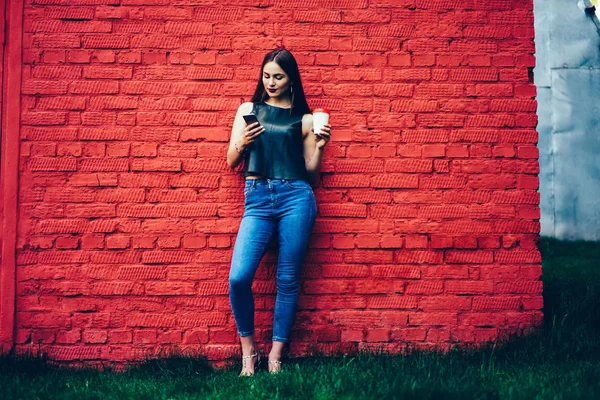 Pensive female student dressed in fashionable outfit enjoying recreation with aroma coffee and sharing news with followers in social network on telephone during standing next to wall with copy space