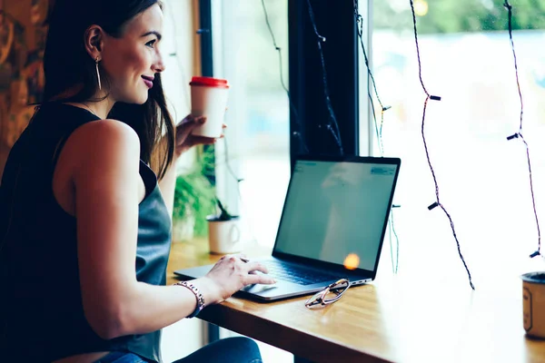 Happy cute blogger dressed in stylish outfit enjoying leisure time with tasty coffee sitting at table with gadget.Positive hipster girl working on laptop computer with mock up screen for your content