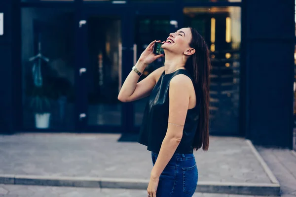 Charming emotional female blogger sincerely laughing while communicating with friends on smartphone connected to internet.Smiling brunette walking at summer street and talking on modern telephone