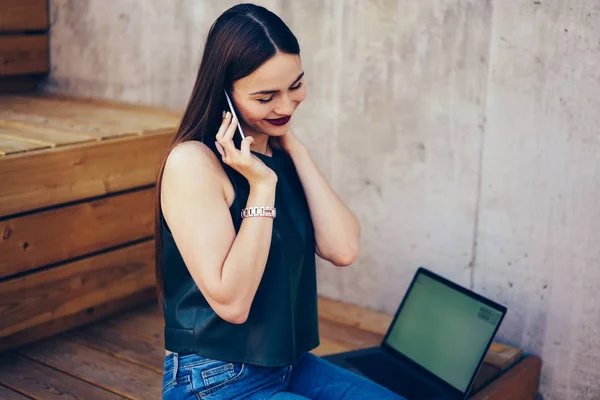 可爱的快乐时髦的女孩享受闲暇时间 而在手机上交谈 坐在靠近笔记本电脑与空白屏幕 积极迷人的黑发在城市环境中再造现代设备 — 图库照片