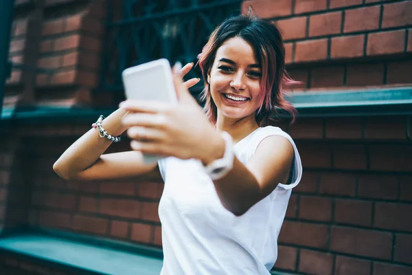 Funny young woman with short haircut making selfie photo on modern smartphone device standing outdoors.Positive hipster girl talking with friend via video chat on telephone during walk in urban setting
