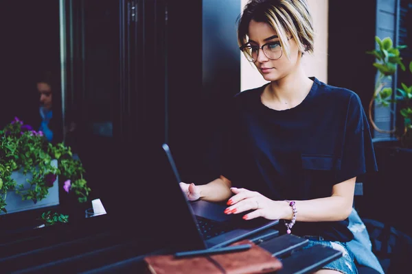Concentrated female graphic designer in stylish eyeglasses working at modern laptop computer sitting in coffee shop interior outdoors.Young woman freelancer typing information on digital netbook