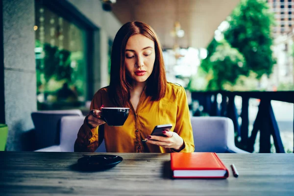 屋外作業の休憩中に香りのコーヒーと近代的なデバイスで再現する豪華な赤毛の女性を熟考します 携帯電話での個人的なウェブサイトで美しい物思いにふける女子学生ブログ — ストック写真