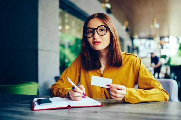 Portrait of charming redhead business woman in stylish eyeglasses looking at camera while holding visit card.Young talented student writing down information to notepad during sitting in coffee shop