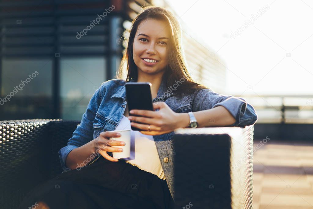 Cheerful hipster girl dressed in denim jacket sending sms message to friend on modern smartphone device.Positive blogger chatting online on mobile phone enjoying tasty coffee sitting outdoors in cafe