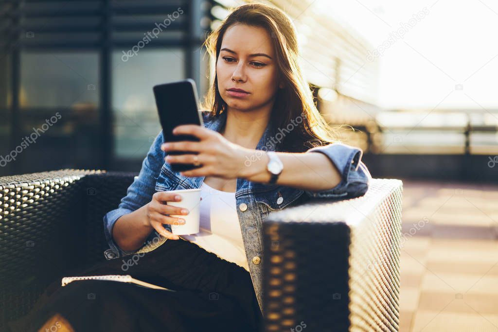 Attractive businesswoman with cup of coffee in hand making internet payment via telephone using high speed 4G connection sitting outdoors in cafeteria.Charming female blogger chatting on cellular