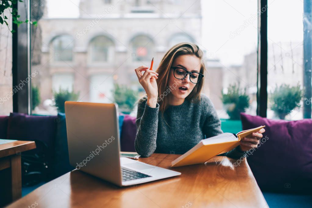 Skilled student in stylish eyeglasses making notes in textbook and preparing for upcoming seminar in coworking space.Clever hipster girl in optical spectacles reading book sitting at laptop computer