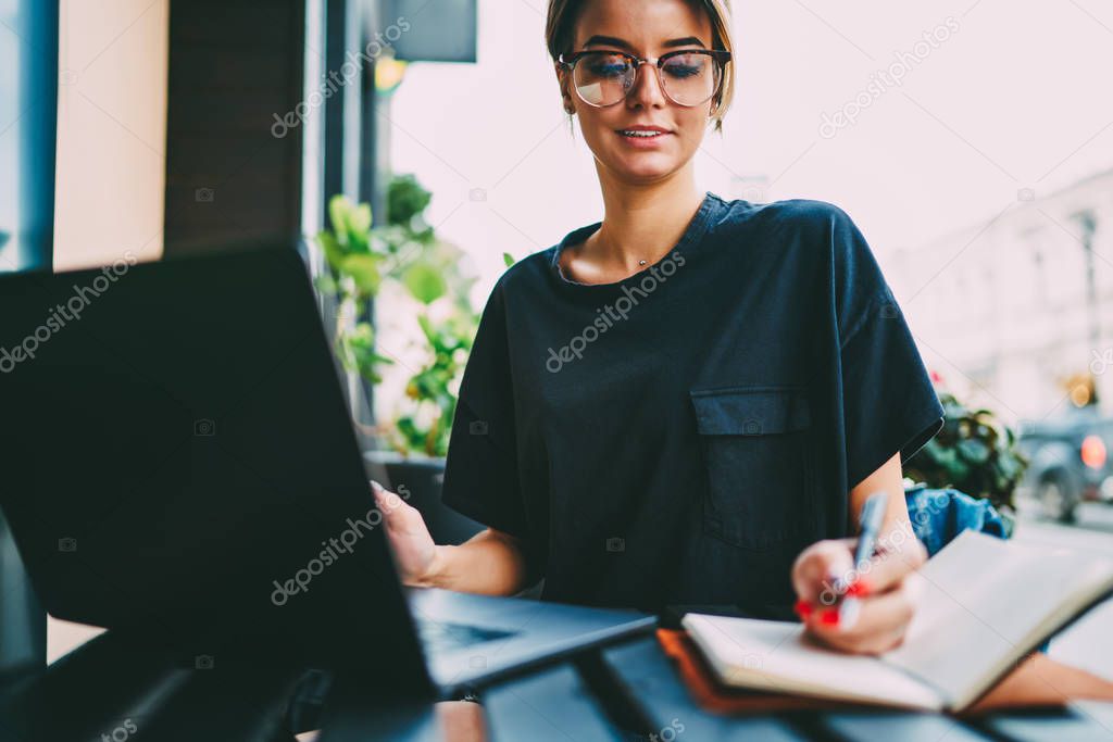 Cropped image of young skilled copywriter in cool eyeglasses searching information in websites and writing down to notepad.Young female entrepreneur recording ideas for business project outside