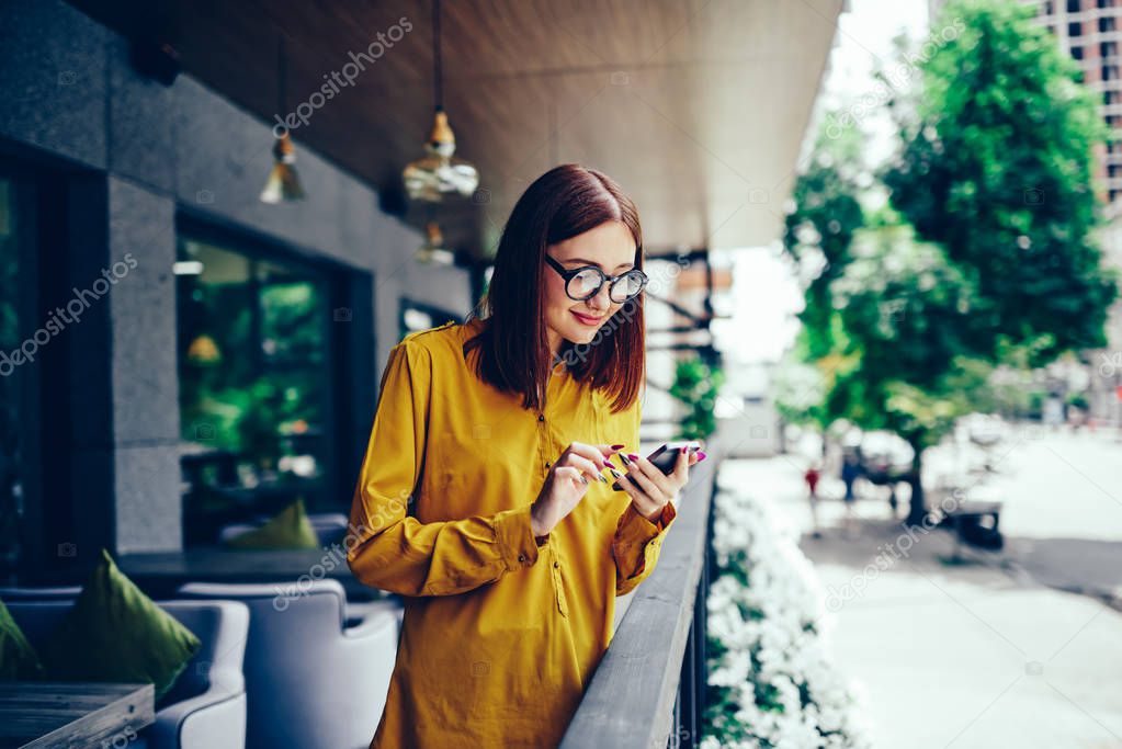 Attractive positive hipster girl in stylish eyewear sending funny messages to friends during recreation time outdoors.Happy cute female reading interesting news in website on modern smartphone