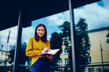 Yarım uzunluk portre sevimli olumlu öğrencinin not defteri zamanlamadan denetlerken kameraya bakarak trendy giyim giymiş. Şirin hippi kız ofis dışında Binası yakınında yürüyüş
