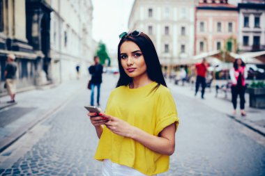 Yarım uzunluk portre sevimli esmer Blogger takipçileri sosyal ağ Haberler paylaşırken kameraya bakıyor. Çekici kız öğrencinin yaz sokak modern cihaz ile gezinme