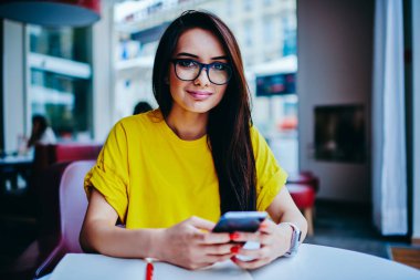 Kameraya bakarak ve kişisel web sitesi içinde takipçileri ile haber ve fikir paylaşımı olumlu esmer blogger yarım uzunluk portre. Modern telefon gözlük manifatura geribildirim şirin hippi kız