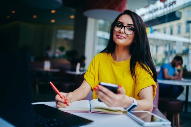 Büyüleyici yaratıcı metin yazarı yarım uzunluk portre rahat giyim için not defteri bilgileri yazma giymiş. Modern cihazlar kullanarak kahve dükkanında çalışan şirin pozitif kadın serbest meslek