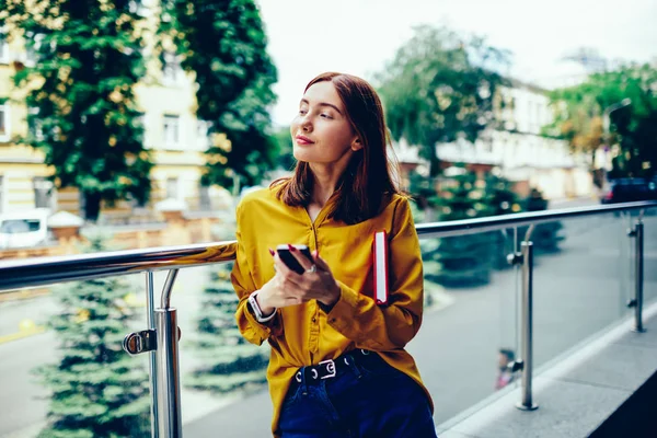 Pondering gorgeous blogger thinking about creative ideas for personal website while walking at urban setting.Positive young woman spending leisure time with modern device during standing outdoors