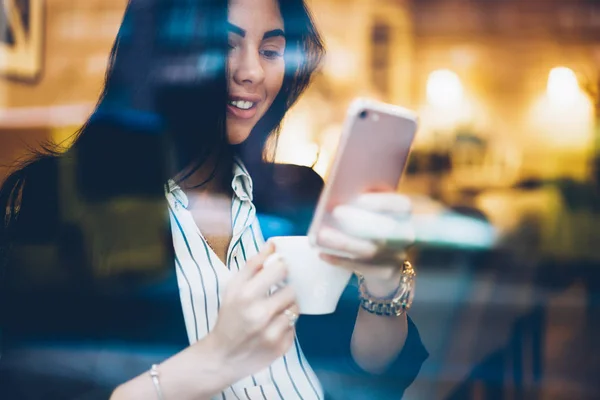 Cropped image of charming positive female person resting in coffee shop with aroma coffee and sending funny messages to friends via application on phone.Young happy woman enjoying leisure time indoors