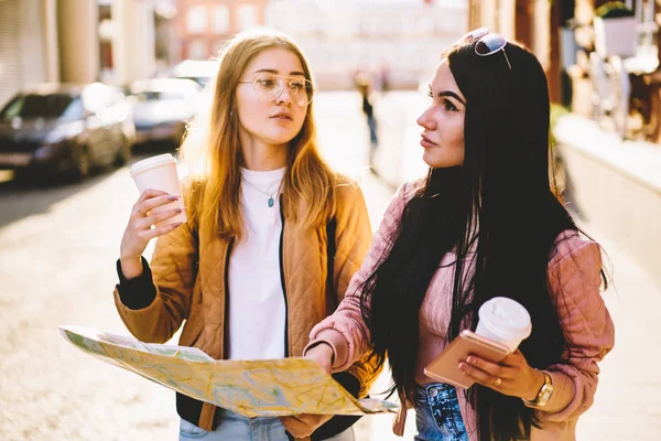 Brunette and blonde female tourist using map for searching route walking with coffee to go during city tour, attractive best friends checking location strolling together in downtown discussing way