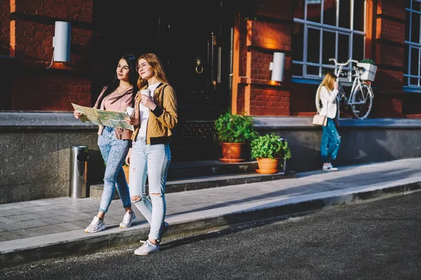 Smiling young women in casual wear walking together on street holding map for getting to location, best friends having city tour together with coffee to go during spring weekends in old town