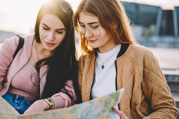 Pensive female travelers reading map together planning route for strolling in downtown,concentrated hipster girls talking about direction to showplaces spending journey together on spring weekends