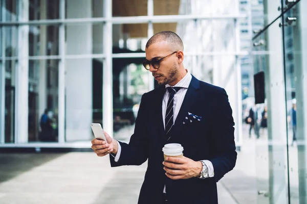 自信的男性企业家在时髦的太阳镜阅读新闻从网络上的移动期间 咖啡休息 自豪的首席执行官在正式的穿着举行外卖咖啡短信信息在智能手机户外 — 图库照片