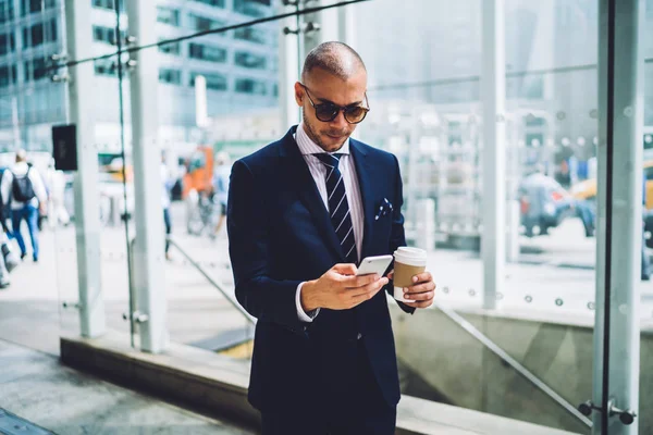 Confident businessman checking email on smartphone during break holding coffee to go, serious male entrepreneur in sunglasses using banking application on mobile for making money transaction outdoor